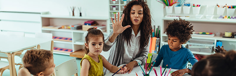 Three young children sitting with a female teacher at a small table within a classroom.