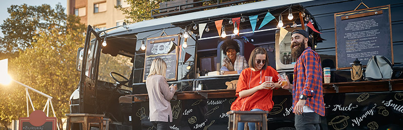 Three customers standing in line outside of a food truck. Owner is serving food from the window.